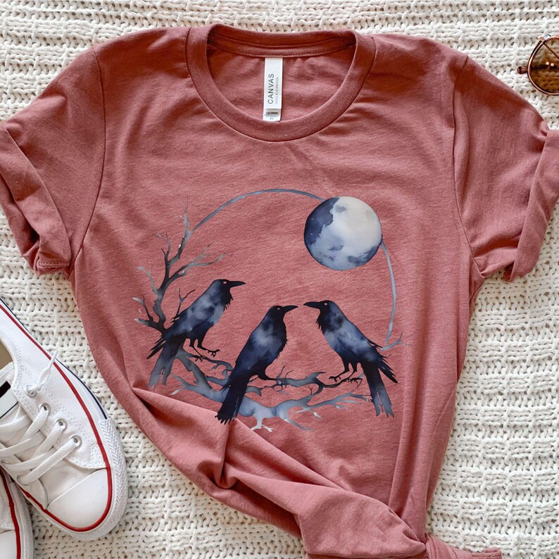 May include: A dusty rose-colored t-shirt featuring a watercolor design of three black crows perched on a branch beneath a full moon. The graphic is enclosed in a circular shape. The shirt has a crew neck and short sleeves.