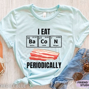 Puede incluir: Camiseta azul claro con el texto negro "I EAT BACON PERIODICALLY". El dise&ntilde;o incluye una ilustraci&oacute;n de tocino y los elementos Ba, Co y N de la tabla peri&oacute;dica. La camiseta es de color azul hielo jaspeado.