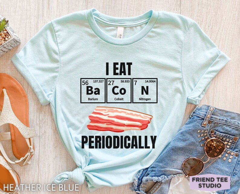 Puede incluir: Camiseta azul claro con el texto negro "I EAT BACON PERIODICALLY". El dise&ntilde;o incluye una ilustraci&oacute;n de tocino y los elementos Ba, Co y N de la tabla peri&oacute;dica. La camiseta es de color azul hielo jaspeado.