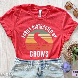 May include: Heather red t-shirt with a vintage-style graphic of a crow with the text "Easily Distracted By Crows".