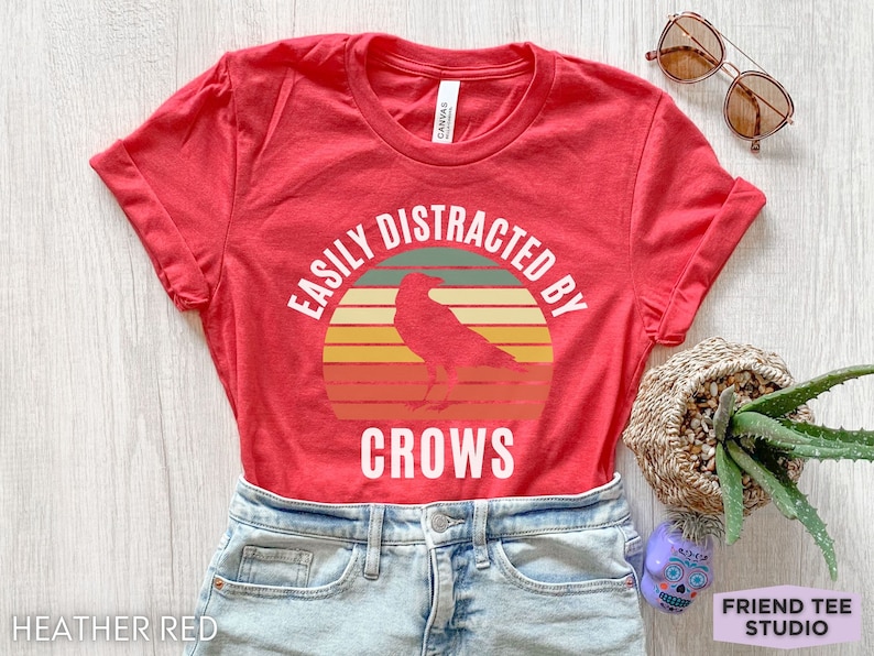 May include: Heather red t-shirt with a vintage-style graphic of a crow with the text "Easily Distracted By Crows".