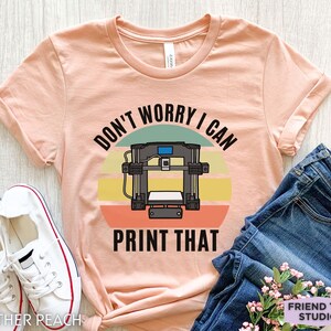 May include: A heather peach t-shirt featuring a graphic of a 3D printer centered over a multi-colored sunset circle. The text "DON'T WORRY I CAN PRINT THAT" is printed in black capital letters above and below the printer illustration.