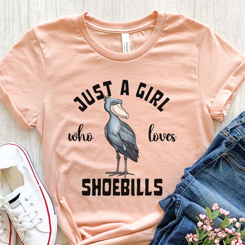 Puede incluir: Una camiseta de color melocot&oacute;n con el texto "Just a girl who loves shoebills" en negro. Un p&aacute;jaro zancudo de dibujos animados se encuentra en el centro del texto.