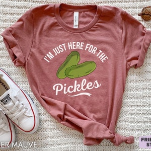May include: Heather mauve t-shirt with the text "I'M JUST HERE FOR THE Pickles" and a graphic of two green pickles. The shirt is paired with white sneakers and sunglasses.