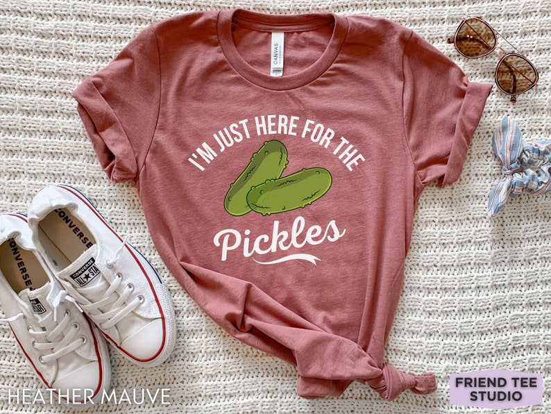 May include: Heather mauve t-shirt with the text "I'M JUST HERE FOR THE Pickles" and a graphic of two green pickles. The shirt is paired with white sneakers and sunglasses.