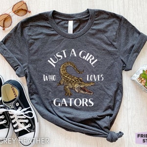 Puede incluir: Camiseta gris oscuro jaspeado con el texto "JUST A GIRL WHO LOVES GATORS" y una ilustraci&oacute;n de un caim&aacute;n. La imagen incluye zapatillas negras, gafas de sol y una peque&ntilde;a planta en maceta. Una prenda informal.