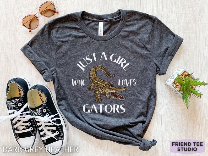 Puede incluir: Camiseta gris oscuro jaspeado con el texto "JUST A GIRL WHO LOVES GATORS" y una ilustraci&oacute;n de un caim&aacute;n. La imagen incluye zapatillas negras, gafas de sol y una peque&ntilde;a planta en maceta. Una prenda informal.