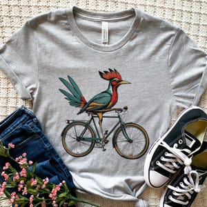 K&ouml;nnte beinhalten: Hellgraues T-Shirt mit einer farbenfrohen Illustration eines Roadrunner-Vogels auf einem Fahrrad. Der Vogel hat rote, blaue und gelbe Federn. Das Fahrrad ist dunkelgr&uuml;n mit beigefarbenen Reifen. Schwarze Leinenschuhe und blaue Jeans sind im Hintergrund.