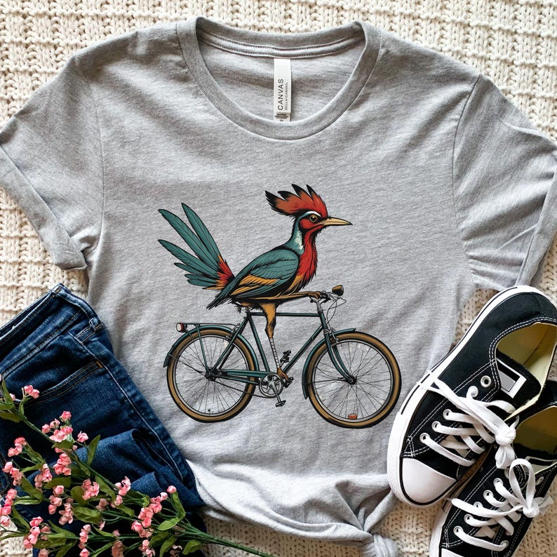K&ouml;nnte beinhalten: Hellgraues T-Shirt mit einer farbenfrohen Illustration eines Roadrunner-Vogels auf einem Fahrrad. Der Vogel hat rote, blaue und gelbe Federn. Das Fahrrad ist dunkelgr&uuml;n mit beigefarbenen Reifen. Schwarze Leinenschuhe und blaue Jeans sind im Hintergrund.