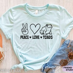 May include: Light blue t-shirt with the text "PEACE LOVE TOADS" and a graphic of a peace sign, heart, and a toad. The shirt is made of a soft material and is tied at the waist. The words "HEATHER ICE BLUE" are at the bottom left.