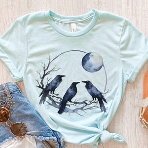 May include: Light blue t-shirt featuring a watercolor design of three black crows perched on a branch, with a full moon in the background. The design is enclosed in a circular shape. The shirt is made of soft material.