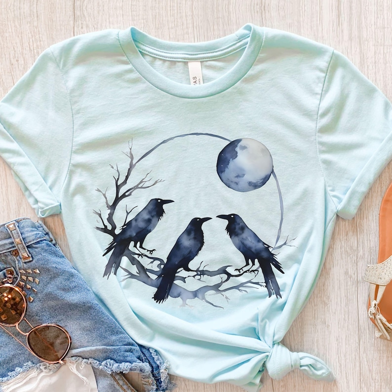 May include: Light blue t-shirt featuring a watercolor design of three black crows perched on a branch, with a full moon in the background. The design is enclosed in a circular shape. The shirt is made of soft material.