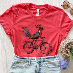K&ouml;nnte beinhalten: Rotes T-Shirt mit einer Illustration eines Vogels mit rotem Kamm und blauen und gr&uuml;nen Federn, der auf einem schwarzen Fahrrad mit braunen Reifen sitzt. Das T-Shirt wird mit Jeansshorts kombiniert. Das T-Shirt ist aus Baumwolle.