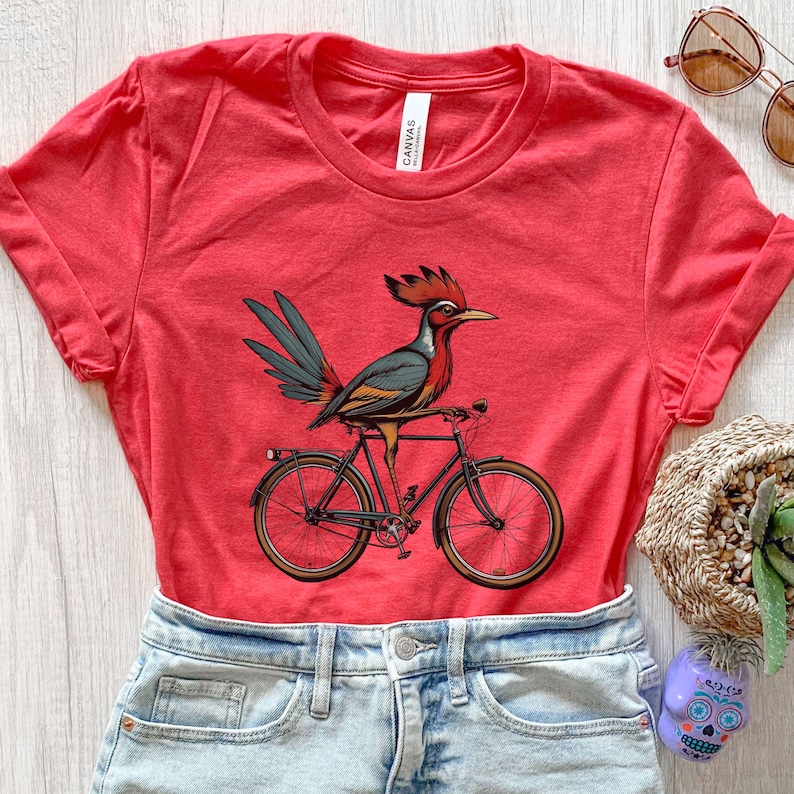 K&ouml;nnte beinhalten: Rotes T-Shirt mit einer Illustration eines Vogels mit rotem Kamm und blauen und gr&uuml;nen Federn, der auf einem schwarzen Fahrrad mit braunen Reifen sitzt. Das T-Shirt wird mit Jeansshorts kombiniert. Das T-Shirt ist aus Baumwolle.