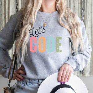 K&ouml;nnte beinhalten: Ein graues Sweatshirt mit dem Text "Let's CODE" in Schwarz, Rosa, Orange, Blau und Gr&uuml;n. Das Sweatshirt hat einen Rundhalsausschnitt. Die Person h&auml;lt einen wei&szlig;en Hut.