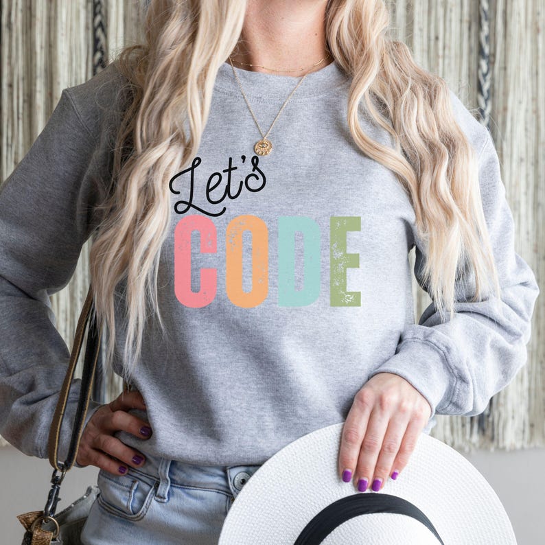 K&ouml;nnte beinhalten: Ein graues Sweatshirt mit dem Text "Let's CODE" in Schwarz, Rosa, Orange, Blau und Gr&uuml;n. Das Sweatshirt hat einen Rundhalsausschnitt. Die Person h&auml;lt einen wei&szlig;en Hut.