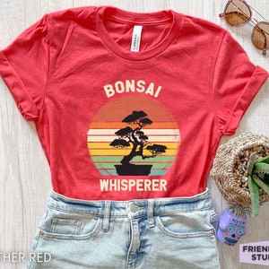 May include: Red t-shirt with a vintage-style graphic of a bonsai tree and the text "Bonsai Whisperer".
