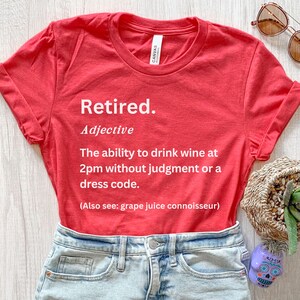 May include: A red t-shirt with the word "Retired" printed in white, along with a definition about drinking wine. The shirt is paired with denim shorts and accessories, including sunglasses and a decorative basket.