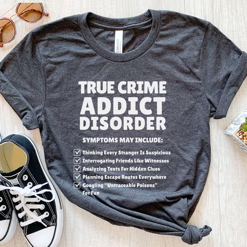 May include: Charcoal grey t-shirt with the white text "TRUE CRIME ADDICT DISORDER". The shirt lists symptoms such as "Thinking Every Stranger Is Suspicious" and "Googling 'Untraceable Poisons' For Fun."