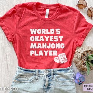 May include: Red t-shirt with white text that reads "World's Okayest Mahjong Player" and a white illustration of a mahjong tile.