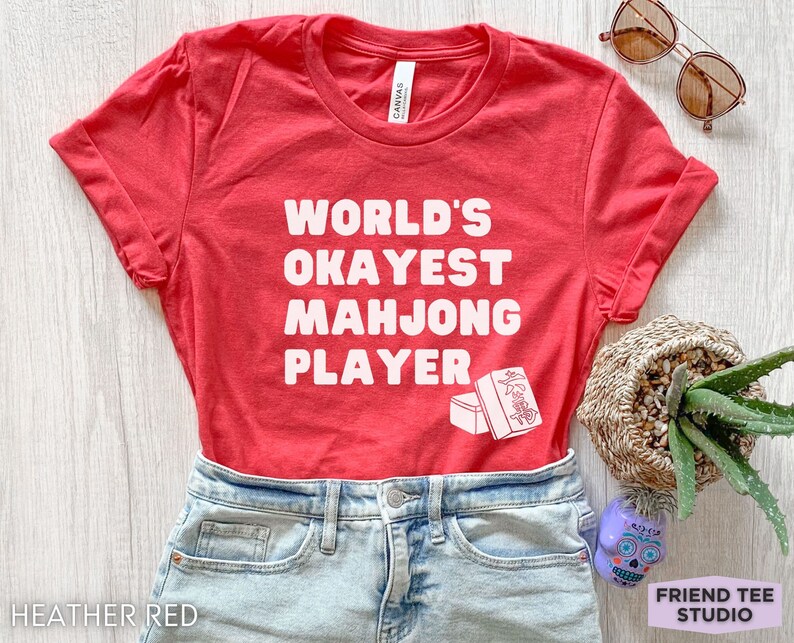 May include: Red t-shirt with white text that reads "World's Okayest Mahjong Player" and a white illustration of a mahjong tile.