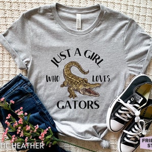 Puede incluir: Camiseta gris jaspeado con el texto "JUST A GIRL WHO LOVES GATORS" y una ilustraci&oacute;n de un caim&aacute;n. La camiseta se combina con vaqueros azules y zapatillas negras. La camiseta es de material atl&eacute;tico.