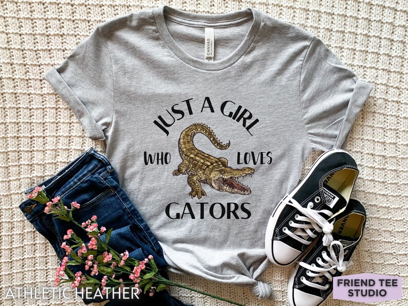 Puede incluir: Camiseta gris jaspeado con el texto "JUST A GIRL WHO LOVES GATORS" y una ilustraci&oacute;n de un caim&aacute;n. La camiseta se combina con vaqueros azules y zapatillas negras. La camiseta es de material atl&eacute;tico.
