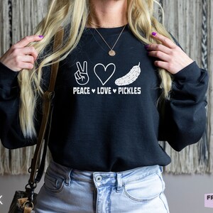 Peut inclure: Un sweat-shirt noir à manches longues avec une impression graphique blanche sur la poitrine. Le motif comprend un signe de paix, un cœur et une illustration de cornichon au-dessus du texte PEACE LOVE PICKLES, avec des petits cœurs séparant les mots.