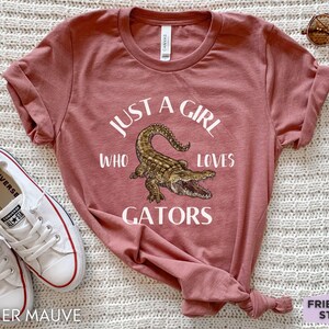 Puede incluir: Camiseta color malva jaspeado con el texto "JUST A GIRL WHO LOVES GATORS" y una ilustraci&oacute;n detallada de un caim&aacute;n. La camiseta se combina con zapatillas blancas y gafas de sol.