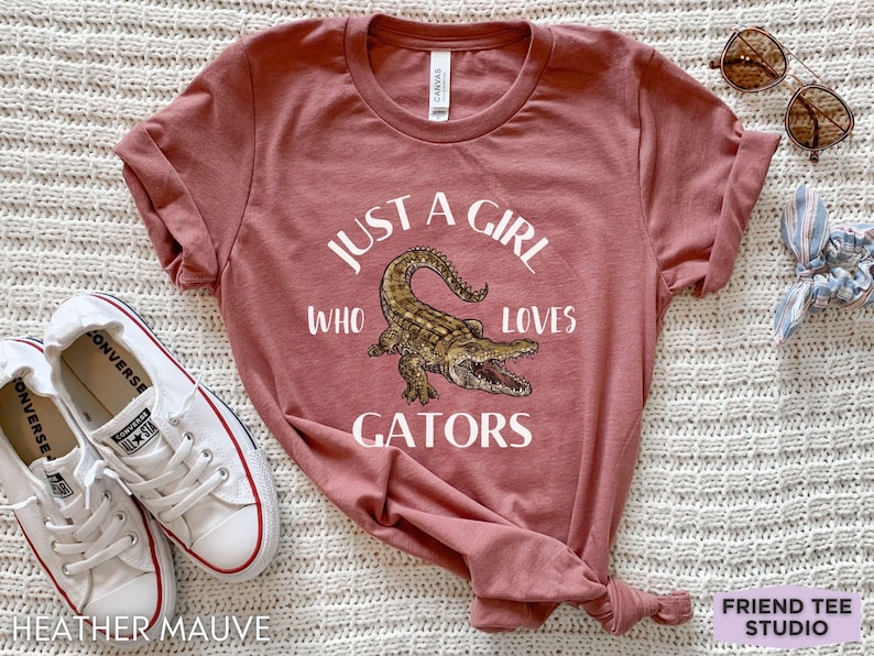Puede incluir: Camiseta color malva jaspeado con el texto "JUST A GIRL WHO LOVES GATORS" y una ilustraci&oacute;n detallada de un caim&aacute;n. La camiseta se combina con zapatillas blancas y gafas de sol.