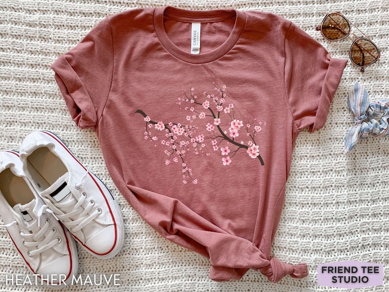 Puede incluir: Camiseta de color malva jaspeado con un gr&aacute;fico de rama de flor de cerezo rosa. La camiseta est&aacute; sobre una superficie blanca y peluda con un par de zapatillas blancas, gafas de sol y una cinta para el pelo azul y blanca.