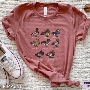 Peut inclure: Un t-shirt mauve chin&eacute; avec une impression graphique de huit esp&egrave;ces de canards diff&eacute;rentes, accompagn&eacute;es de leurs noms : Mallard, Wood, Baikal Teal, Gadwall, Eurasian Teal, Redhead, Rosy-billed Pochard et Red-crested Pochard.