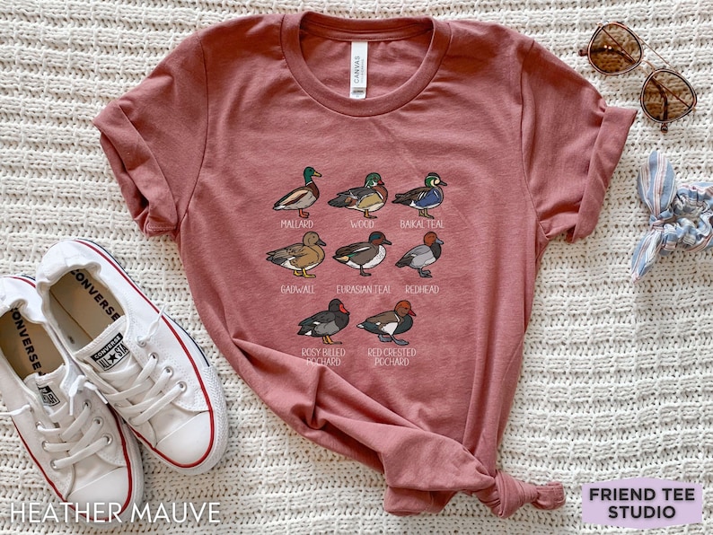 Peut inclure: Un t-shirt mauve chin&eacute; avec une impression graphique de huit esp&egrave;ces de canards diff&eacute;rentes, accompagn&eacute;es de leurs noms : Mallard, Wood, Baikal Teal, Gadwall, Eurasian Teal, Redhead, Rosy-billed Pochard et Red-crested Pochard.