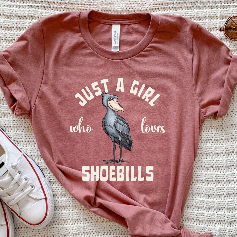 Puede incluir: Una camiseta de color marr&oacute;n rojizo con el texto "Just a girl who loves shoebills" y una ilustraci&oacute;n de dibujos animados de una cig&uuml;e&ntilde;a zancuda.