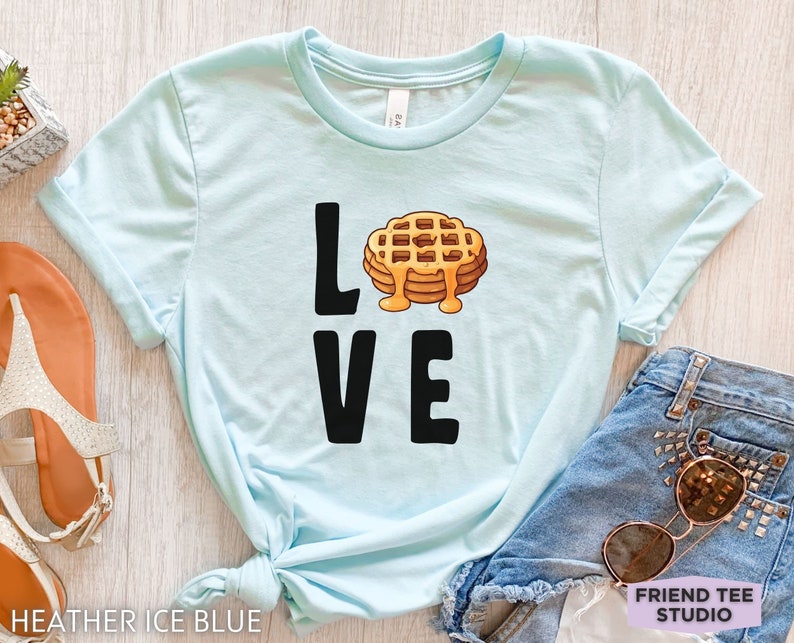 K&ouml;nnte beinhalten: Hellblaues T-Shirt mit dem Wort "LOVE" in schwarzen Buchstaben und einer Waffelgrafik in der Mitte des Wortes. Die Waffel ist gelb und braun mit einem Schuss Sirup.