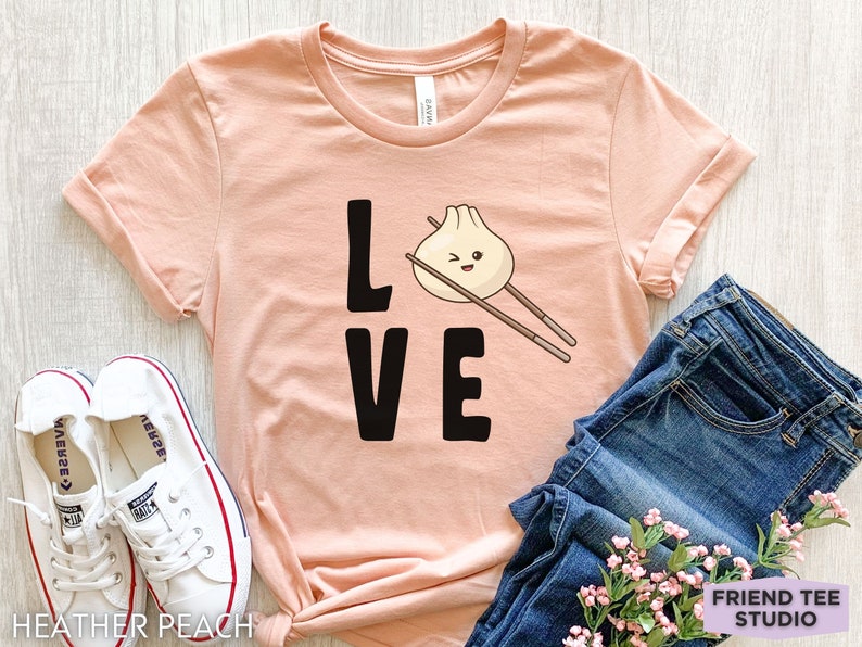 Op de afbeelding: Een perzikkleurig T-shirt met het woord "LOVE" in zwart gedrukt. De "O" in LOVE is vervangen door een cartoon-dumpling met eetstokjes.