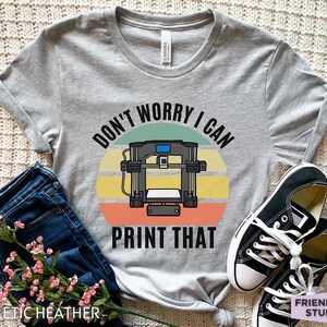May include: A heather gray short-sleeve t-shirt featuring a graphic of a 3D printer centered over a multi-colored sunset circle. The text reads "DON'T WORRY I CAN PRINT THAT" in black capital letters. The shirt has a crew neckline and rolled sleeves.