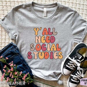 May include: A gray t-shirt with the text "Y'all Need Social Studies" in a retro, colorful font.