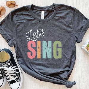 Op de afbeelding: Donkergrijs t-shirt met de woorden "Let's SING" in witte letters en kleurrijke blokletters. Het woord "SING" is roze, oranje, turquoise en groen. Zwart-witte sneakers en een zonnebril zijn zichtbaar.