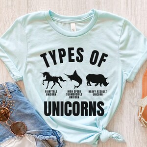 May include: A light blue t-shirt with the text "Types of Unicorns" printed in black. The shirt features three black silhouettes of different unicorns: a fairytale unicorn, a high-speed submersible unicorn, and a heavy assault unicorn.