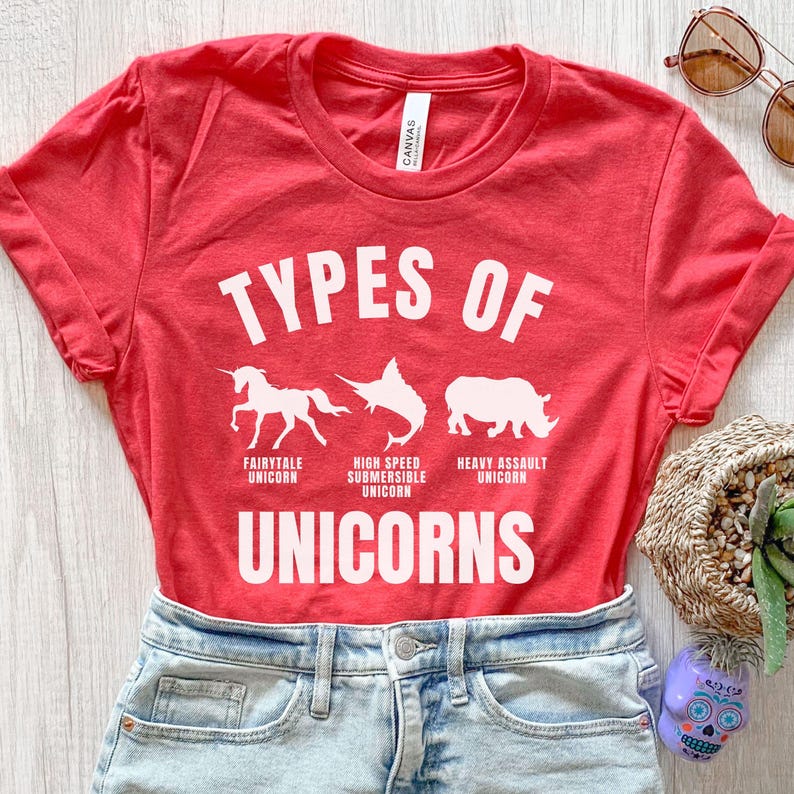 May include: Red t-shirt with white text that reads "Types of Unicorns". The shirt features three white silhouettes of a unicorn, a swordfish, and a rhinoceros. The text below each silhouette reads "Fairytale Unicorn", "High Speed Submersible Unicorn", and "Heavy Assault Unicorn".