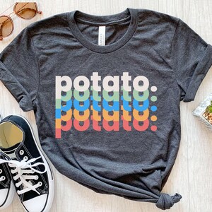 May include: A dark heather gray short-sleeve t-shirt featuring the word potato printed four times in a stacked, overlapping design. The text appears in white, light green, blue, and orange, with each line slightly offset from the one above.