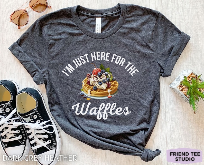 Op de afbeelding: Donkergrijs heather t-shirt met de tekst "I'm just here for the waffles" in wit. Een cartoonwafel met slagroom, blauwe bessen en aardbeien is afgebeeld boven de tekst.