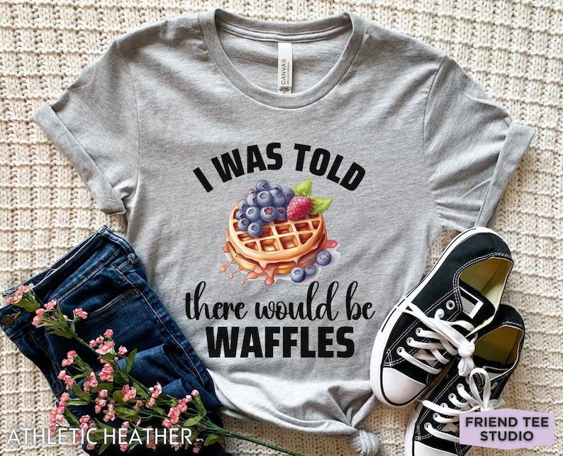 May include: Athletic heather gray t-shirt with the text "I WAS TOLD there would be WAFFLES". The graphic features a waffle topped with blueberries and a raspberry. A pair of black canvas sneakers and blue jeans are next to the shirt.