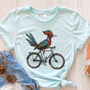 K&ouml;nnte beinhalten: Hellblaues T-Shirt mit einem bunten Vogel auf einem Fahrrad. Der Vogel hat rote, teal- und gelbe Federn mit einem roten Kamm. Das Fahrrad ist schwarz mit braunen Reifen. Das Shirt ist aus weichem Material.