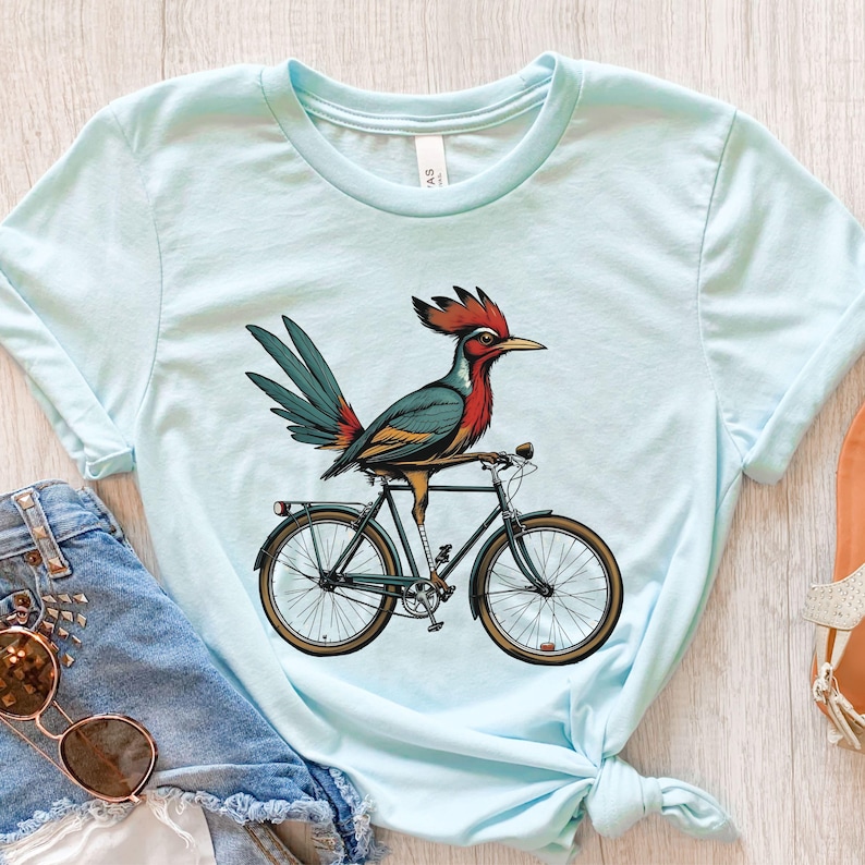 K&ouml;nnte beinhalten: Hellblaues T-Shirt mit einem bunten Vogel auf einem Fahrrad. Der Vogel hat rote, teal- und gelbe Federn mit einem roten Kamm. Das Fahrrad ist schwarz mit braunen Reifen. Das Shirt ist aus weichem Material.
