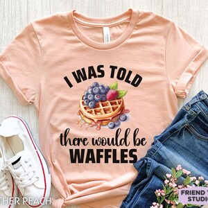 May include: A peach-colored t-shirt with the text "I WAS TOLD there would be WAFFLES" in black. The graphic features a waffle topped with blueberries and a raspberry. White sneakers and blue jeans are also in the image.