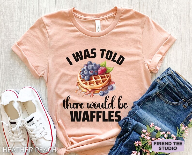 May include: A peach-colored t-shirt with the text "I WAS TOLD there would be WAFFLES" in black. The graphic features a waffle topped with blueberries and a raspberry. White sneakers and blue jeans are also in the image.