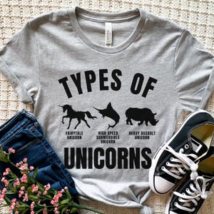 May include: A gray t-shirt with the text "Types of Unicorns" printed on it. The shirt features three black silhouettes of unicorns: a fairytale unicorn, a high-speed submersible unicorn, and a heavy assault unicorn.