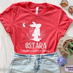Puede incluir: Una camiseta de color rojo jaspeado con un gr&aacute;fico blanco de un conejo, un sol y elementos florales. El texto dice OSTARA seguido de BALANCE, REBIRTH y RENEWAL. La camiseta se combina con vaqueros azul claro, gafas de sol y una suculenta.